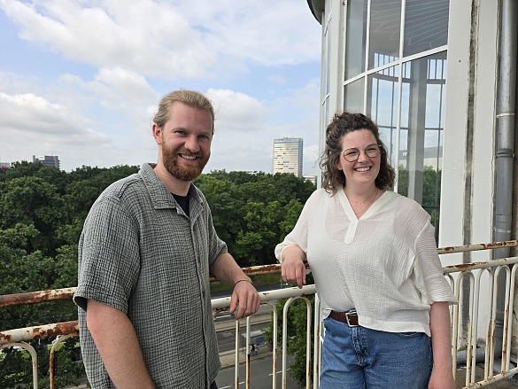 Eric Nissen trägt ein graugrünes, kurzärmeliges Hemd aus Musselinstoff, darunter ein schwarzes T-Shirt. Er trägt einen Bart und seine blonden Haare zu einem Zopf gebunden. Sophie ridder trägt eine blaue Jeans und eine cremefarbene Bluse aus Musselinstoff. Ihre schulterlangen braunen Haaare trägt sie halboffen, auf der Nase sitzt eine schwarzgerahmte Brille. Beide Personen lächeln, während sie auf einem Balkon an einem Geländer am Standort Doventorscontrescarpe stehen.