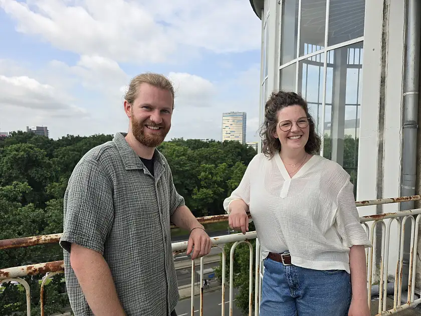Eric Nissen trägt ein graugrünes, kurzärmeliges Hemd aus Musselinstoff, darunter ein schwarzes T-Shirt. Er trägt einen Bart und seine blonden Haare zu einem Zopf gebunden. Sophie ridder trägt eine blaue Jeans und eine cremefarbene Bluse aus Musselinstoff. Ihre schulterlangen braunen Haaare trägt sie halboffen, auf der Nase sitzt eine schwarzgerahmte Brille. Beide Personen lächeln, während sie auf einem Balkon an einem Geländer am Standort Doventorscontrescarpe stehen.