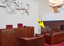 Auf dem Foto ist der Sitzungssaal der Bremischen Bürgerschaft zu sehen. Ein Pfeil zeigt auf den Arbeitsplatz der drei.