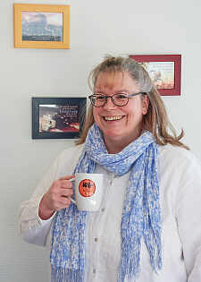 Yvonne Burke hält eine Tasse mit einem Button mit der Aufschrift Wir für Bremen in der Hand, im Hintergrund sind Bilder zu erkennen, die sie aufgehängt hat.