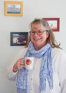 Yvonne Burke hält eine Tasse mit einem Button mit der Aufschrift Wir für Bremen in der Hand, im Hintergrund sind Bilder zu erkennen, die sie aufgehängt hat.