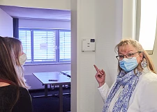 Yvonne Burke zeigt in einen Raum, in dem schon Möbel für neue Kolleg:innen stehen, das Türschild des Raumes ist noch nicht mit den neuen Namen bedruckt.