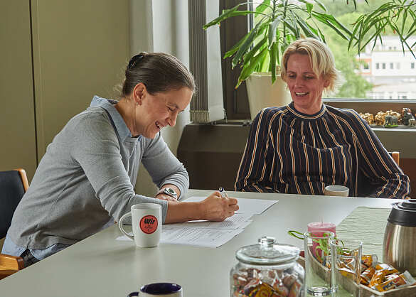 Die zwei Kolleginnen sitzen am Tisch und machen sich lächelnd Notizen