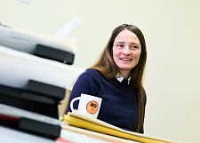 Lachende Frau mit einem dunkelblauen Pullover, langen braunen Haaren am Schreibtisch mit einer Kaffeetasse
