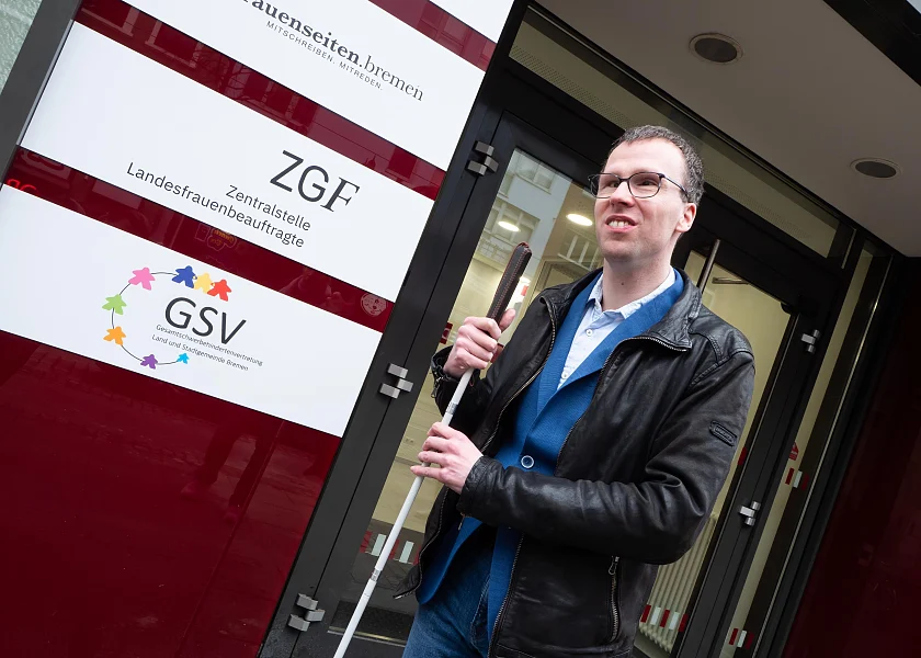 Christian Dabs steht vor einem Gebäude und hält seinen Blindenstock in beiden Händen. Hinter ihm sind Informationstafeln am Gebäude zu sehen.