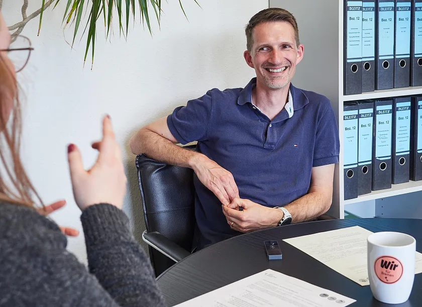 Dennis Kemmerling sitzt in einem Büro an einem Tisch und lächelt, er trägt ein T-Shirt.
