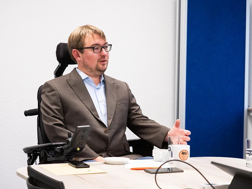 Arne Frankenstein sitzt in seinem elektrischen Rollstuhl an einem Tisch, vor ihm lieben Papierdokumente sowie eine Kaffeetasse und ein Wasserglas. Er gestikuliert im Gespräch mit seiner Hand.