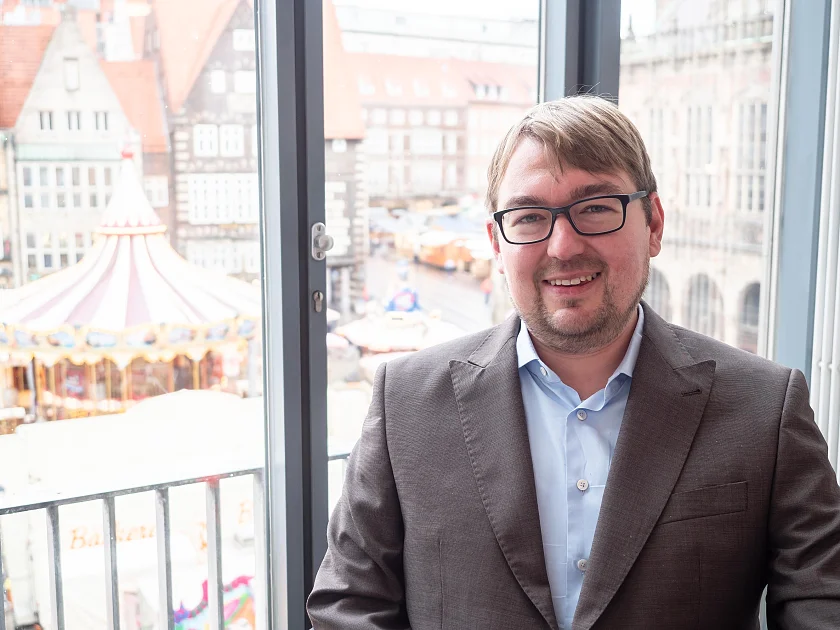 Arne Frankenstein sitzt vor einem Fenster im Festsaal der Bremischen Bürgerschaft, hinter ihm ist der Bremer Marktplatz zu erkennen, auf dem ein historisches Karussell steht.