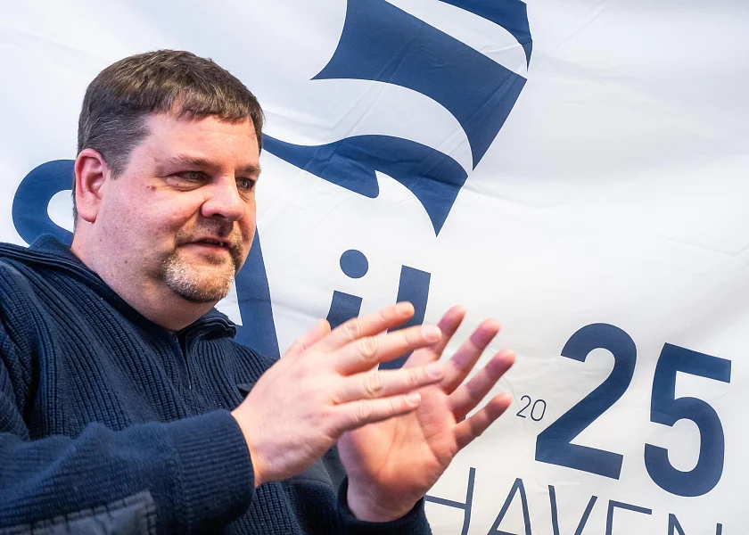 Björn Greifenberger sitzt vor einer Wandflagge der SAIL BREMERHAVEN 2025 und gestikuliert mit den Händen.