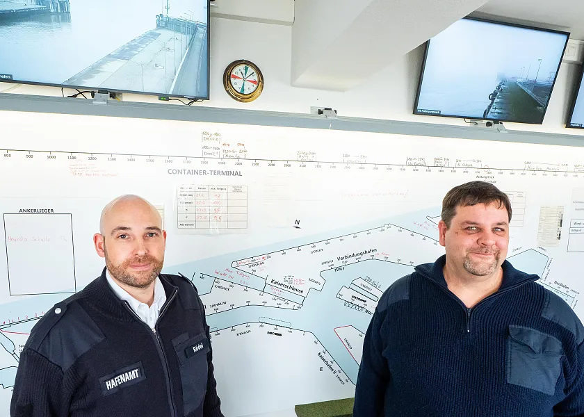 Nils Rödel (links) trägt eine Glatze und seine dunkelblaue Dienstjacke mit dem Patch Hafenamt auf der Brust. Neben ihm steht Björn Greifenberger (rechts), der einen dunkelblauen Strickpullover trägt und lächelt. Hinter den Männern ist ein größerer Ausschnitt der Hafenlandkarte zu erkennen. Über der Karte hängen Monitore, die verschiedene Schleusen des Hafens zeigen.