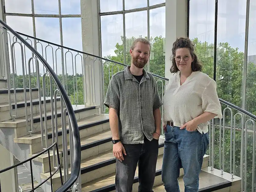 Eric Nissen trägt ein graugrünes, kurzärmeliges Hemd aus Musselinstoff, darunter ein schwarzes T-Shirt. Er trägt einen Bart und seine blonden Haare zu einem Zopf gebunden. Sophie ridder trägt eine blaue Jeans und eine cremefarbene Bluse aus Musselinstoff. Ihre schulterlangen braunen Haaare trägt sie halboffen, auf der Nase sitzt eine schwarzgerahmte Brille. Beide Personen lächeln, während sie in einem verglasten Rundtreppenaufgang an der Doventorscontrescarpe stehen.