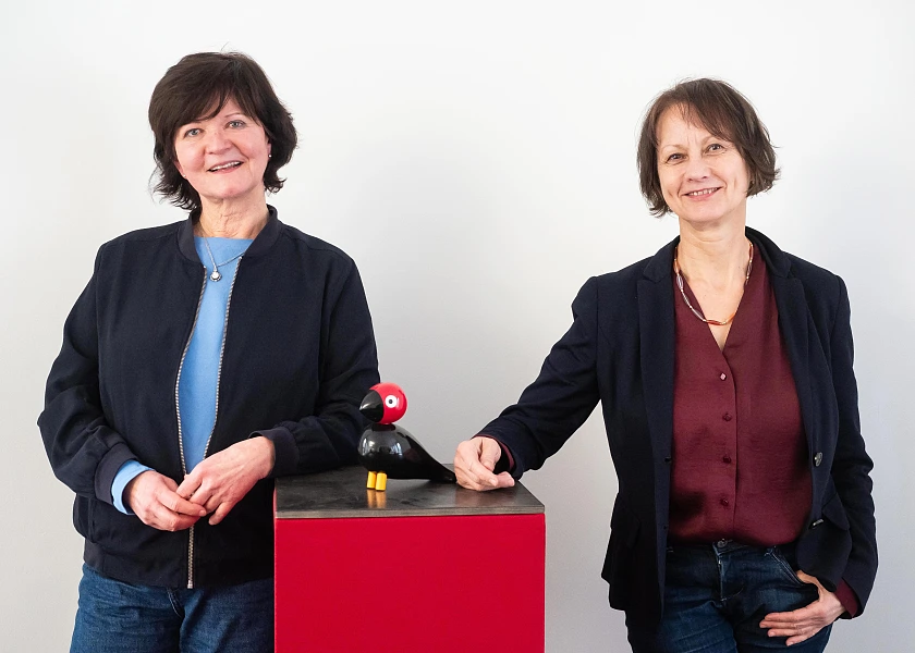 Das Bild zeigt Birgit Sprecher und Karen Rohlf-Grimm, die an einem Tisch stehen, auf dem sich schwarze Vogelfigur mit rotem Kopf befindet.
