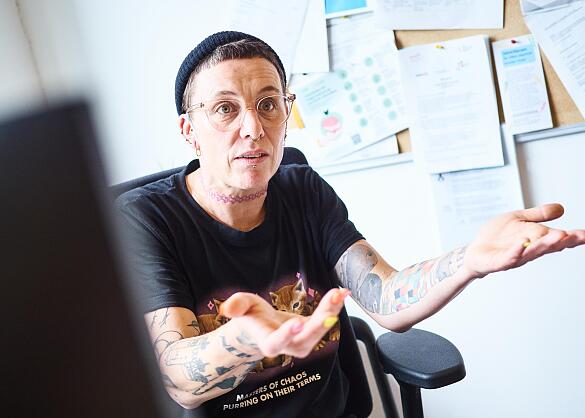 Melanie Dauelsberg sitzt auf Ihrem Bürostuhl und gestikuliert wild mit ihren Händen, während sie von ihrer Arbeit erzählt. Sie trägt kurze Haare, eine Strickmütze, eine Brille und eine rosa Choker-Halskette. Melanie Dauelsberg sitzt auf Ihrem Bürostuhl und gestikuliert wild mit ihren Händen, während sie von ihrer Arbeit erzählt. Sie trägt kurze Haare, eine Strickmütze, eine Brille und eine rosa Choker-Halskette.