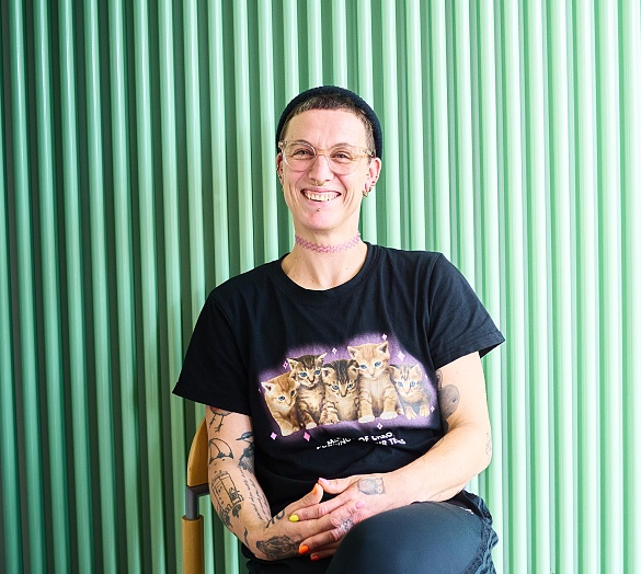 Melanie Dauelsberg sitzt mit überschlagenen Beinen auf einem Stuhl vor einem türkisfarbenen Heizkörper und lächelt in die Kamera. Sie trägt kurze Haare, eine Strickmütze, eine Brille und eine rosa Choker-Halskette. Melanie Dauelsberg sitzt mit überschlagenen Beinen auf einem Stuhl vor einem türkisfarbenen Heizkörper und lächelt in die Kamera. Sie trägt kurze Haare, eine Strickmütze, eine Brille und eine rosa Choker-Halskette.