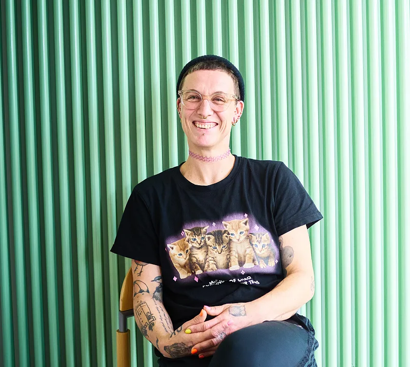 Melanie Dauelsberg sitzt mit überschlagenen Beinen auf einem Stuhl vor einem türkisfarbenen Heizkörper und lächelt in die Kamera. Sie trägt kurze Haare, eine Strickmütze, eine Brille und eine rosa Choker-Halskette.