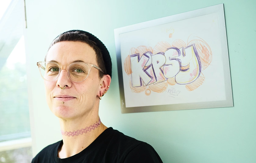 Melanie Dauelsberg steht neben ihrem dienstlichen Briefkasten mit der gemalten Aufschrift KIPSY. Sie trägt kurze Haare, eine Strickmütze, eine Brille und eine rosa Choker-Halskette.