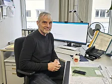 Mann mit grauen Haaren sitzt an seinem Büroarbeitsplatz vor dem Computer, vor ihm auf dem Schreibtisch liegt ein Mobiltelefon