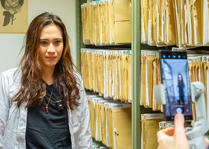 Ina Bloß hat lange braune Haare, die sie offen trägt und steht in dunklem T-Shirt und mit hellgrauer Jacke von Aktenschränken und blickt in eine Smartphone-Kamera.