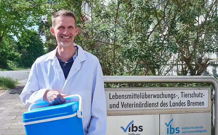 Dennis Kemmerling steht mit weißem Kittel und blauer Kühlbox vor dem Eingangsschild des LMTVet