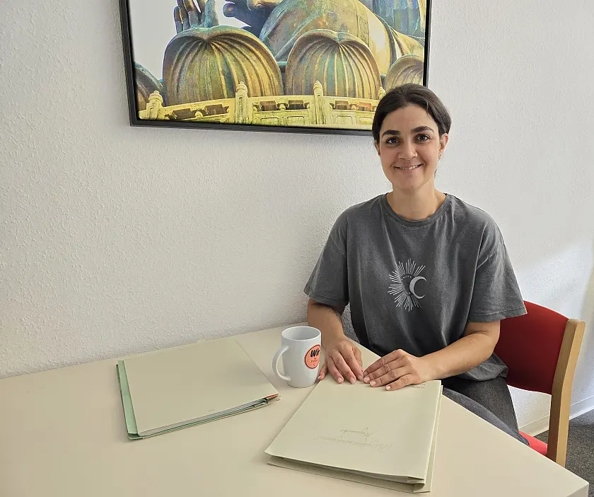 Junge frau mit grauen T-Shirt sitzt an einem Tisch und hat eine Mappe vor sich liegen.