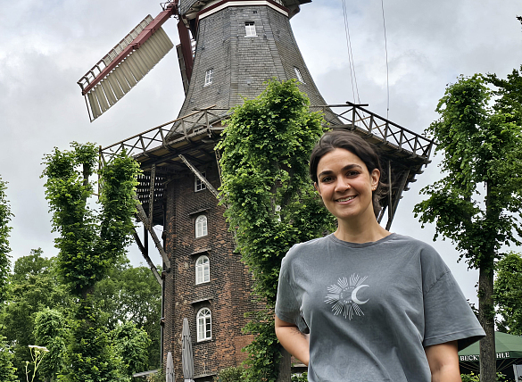 Frau mit grauem T-Shirt und zurückgebundenen, dunklen Haaren steht vor der Mühle am Wall.