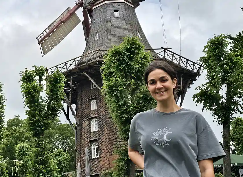 Frau mit grauem T-Shirt und zurückgebundenen, dunklen Haaren steht vor der Mühle am Wall.