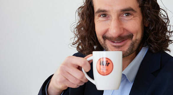 Aykut Tasan lächelt in die Kamera. In der Hand hält er einen Kaffeebecher mit dem Slogan Wir für Bremen.