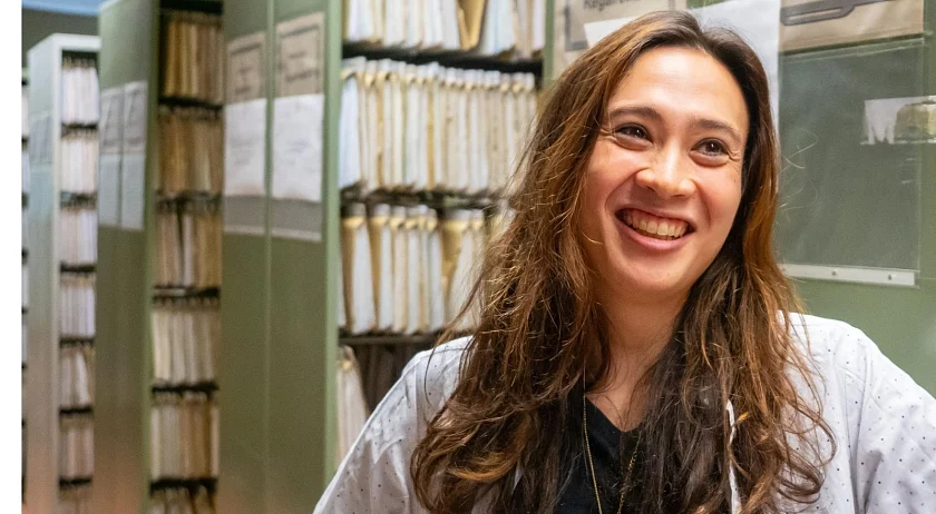 Ina Bloß hat lange braune Haare, die sie offen trägt und steht in dunklem T-Shirt und mit hellgrauer Jacke von Aktenschränken und lächelt.