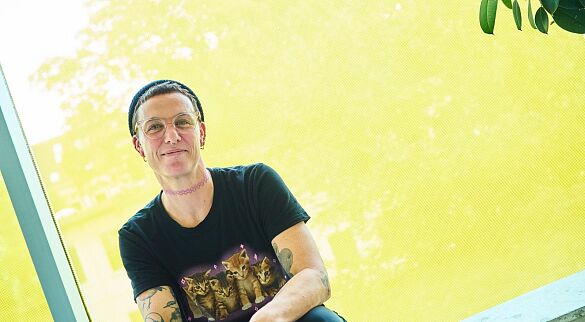 Melanie Dauelsberg sitzt mit einem schwarzen T-Shirt mit Katzenprint und einer Strickmütze vor einem gelb folierten Fenster. Sie trägt außerdem kurz rasierte Haare, eine Brille und eine rosafarbene Choker-Halskette. Melanie Dauelsberg sitzt mit einem schwarzen T-Shirt mit Katzenprint und einer Strickmütze vor einem gelb folierten Fenster. Sie trägt außerdem kurz rasierte Haare, eine Brille und eine rosafarbene Choker-Halskette.