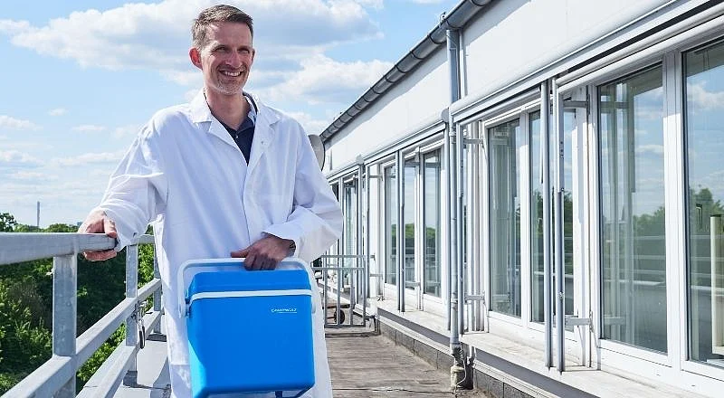 Dennis Kemmerling steht mit einem weißen Kittel und einer kleinen blauen Kühltruhe auf einer Dachterasse.