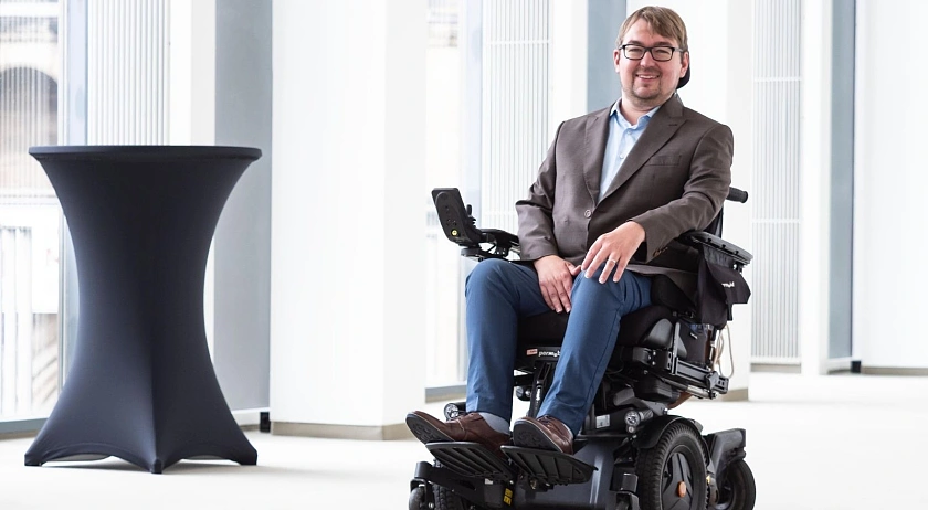 Arne Frankenstein trägt ein braun-graues Sakko und eine blaue Jeans. Er sitzt in einem elektrischen Rollstuhl in einem Flur im Haus der Bürgerschaft.