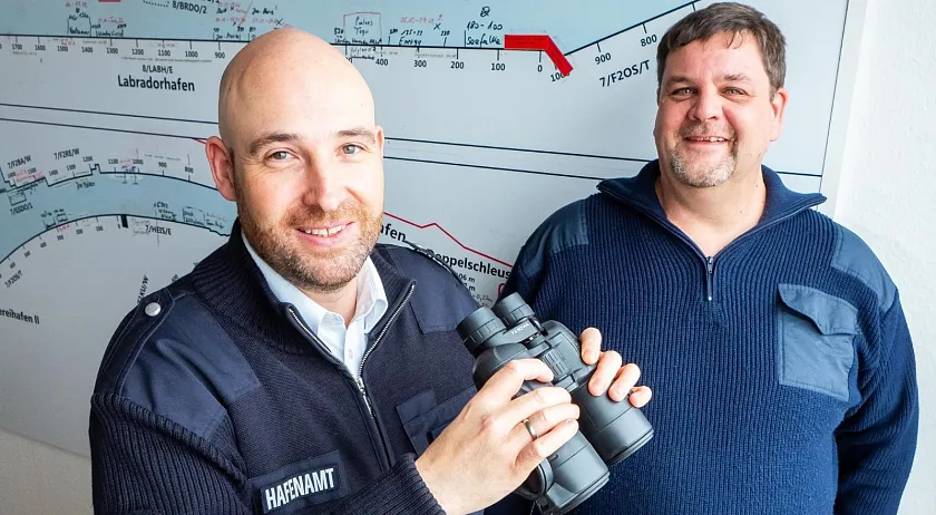 Nils Rödel (links) trägt eine Glatze und seine dunkelblaue Dienstjacke mit dem Patch Hafenamt auf der Brust. In der Hand hält er ein Fernglas. Neben ihm steht Björn Greifenberger (rechts), der einen dunkelblauen Strickpullover trägt und lächelt. Hinter den Männern ist ein Ausschnitt der Hafenlandkarte zu erkennen.