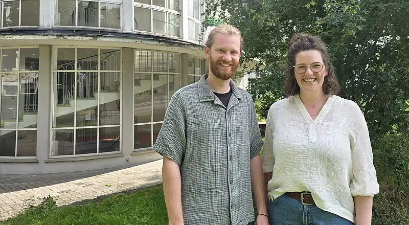 Eric Nissen trägt ein graugrünes, kurzärmeliges Hemd aus Musselinstoff, darunter ein schwarzes T-Shirt. Er trägt einen Bart und seine blonden Haare zu einem Zopf gebunden. Sophie ridder trägt eine blaue Jeans und eine cremefarbene Bluse aus Musselinstoff. Ihre schulterlangen braunen Haaare trägt sie halboffen, auf der Nase sitzt eine schwarzgerahmte Brille. Beide Personen lächeln, während sie von einem verglasten Treppenaufgang an der Doventorscontrescarpe stehen.