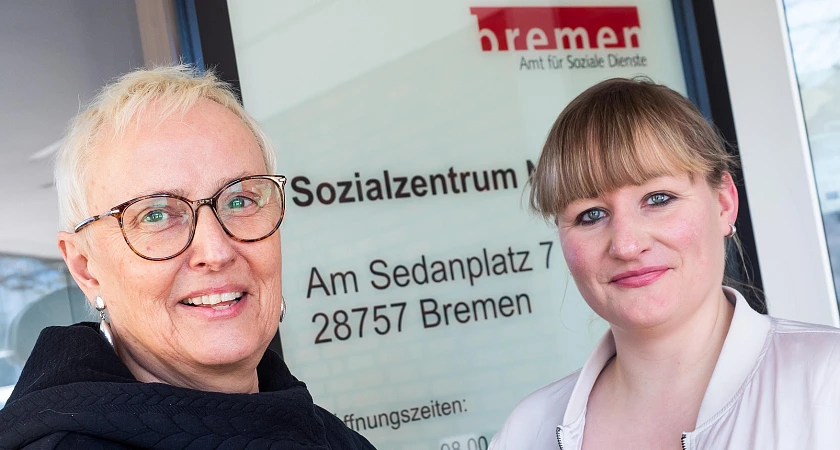 Die blonde, kurzhaarige, lebensältere Verena Stolczewski steht neben ihrer jungen Kollegin Madlen Loske vor dem Eingang des Amts für Soziale Dienste in Bremen Nord, auf der Scheibe hinter ihnen steht Sozialzentrum geschrieben.