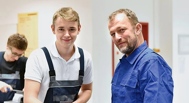 Meister im blauen Overall mit seinem Lehrling in einer Werkstatt, beide schauen in die Kamera