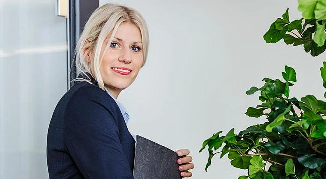 Frau in förmlicher Kleidung, die über die Schulter in die Kamera blickt, sie hält einen Aktenordner in der Hand und geht Richtung Tür.