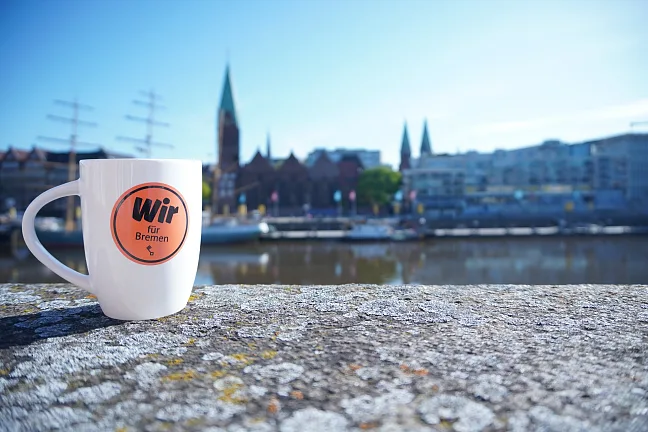 Kaffeetasse mit dem Text Wir für Bremen auf einer Mauer, im Hintergrund sind ein Fluss und Schiffe zu erkennen