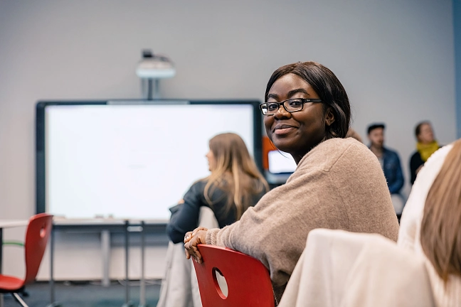 Schwarze Frau, die in einem Studienraum sitzt, hinter ihr ist ein Smartboard zu sehen