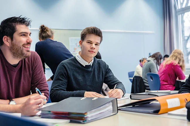 Zwei Schüler in einem Unterrichtsraum mit Ordnern auf dem Tisch