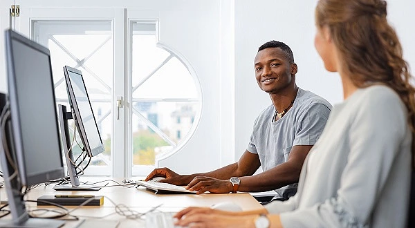 Schwarzer Mann mit kurzen Haaren und grauem T-Shirt sitzt am Schreibtisch vor einem Monitor