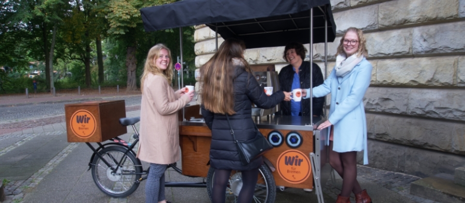 Ein Kaffeefahrrad steht vor dem Haus des Reichs. Es ist beklebt mit runden Buttons mit der Aufschrift "Wir für Bremen" und dem Bremer Schlüssel. Drumherum stehen vier Personen. Drei Personen der Leitstelle Arbeitgeberattraktivität und Alena Baumgarten, die von ihrer Tätigkeit im Interview berichtet.