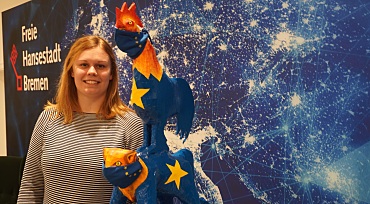 Europablauer Hintergrund mit Sternen, davor steht Lisa Frohböse zwischen dem Logo der Freien Hansestadt Bremen und den Bremer Stadtmusikanten