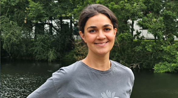 Junge dunkelhaarige Frau mit weggebundenen Haaren stht vor einem Gewässer, im Hintergrund sieht man Bäum. 