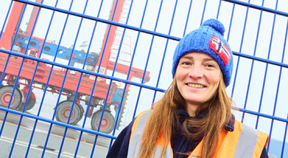 Frau mit orangefarbener Warnweste und blauer Pudelmütze steht vor einem blauen Doppelstabmattenzaun