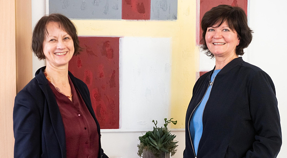 Das Bild zeigt Karen Rohlf-Grimm und Birgit Sprecher, die an einem Tisch stehen, auf dem sich eine Pflanze befindet.