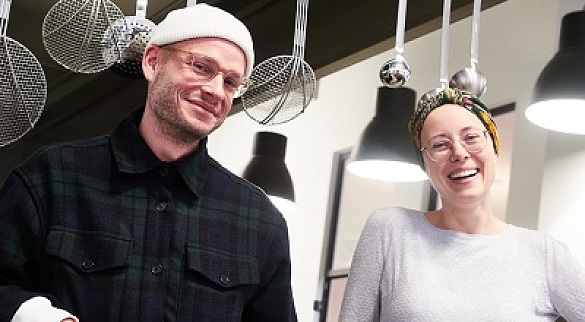 Mann mit dunkler, karierter Jacke, Brille und weißer Mütze steht in einer Großküche neben einer Frau im grauen Pullover, mit Brille und buntem Turban. Hinter Ihnen hängen Suppen- und Schaumkellen.