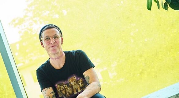 Eine Frau mit ganz kurzen Haaren und eine Strickmütze auf dem Kopf sitzt vor einer gelben Scheibe. Sie trägt eine Brille und ein schwarzes T-Shirt mit Katzen darauf und lächelt in die Kamera.