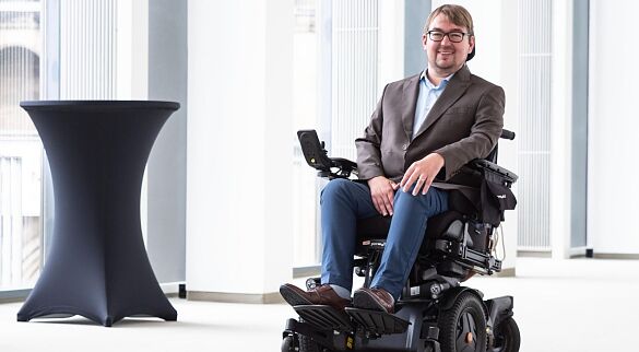 Arne Frankenstein trägt ein braun-graues Sakko und eine blaue Jeans. Er sitzt in einem elektrischen Rollstuhl in einem Flur im Haus der Bürgerschaft.