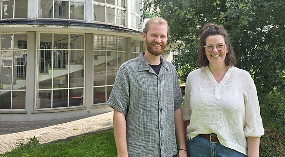 Eric Nissen trägt ein graugrünes, kurzärmeliges Hemd aus Musselinstoff, darunter ein schwarzes T-Shirt. Er trägt einen Bart und seine blonden Haare zu einem Zopf gebunden. Sophie ridder trägt eine blaue Jeans und eine cremefarbene Bluse aus Musselinstoff. Ihre schulterlangen braunen Haaare trägt sie halboffen, auf der Nase sitzt eine schwarzgerahmte Brille. Beide Personen lächeln, während sie von einem verglasten Treppenaufgang an der Doventorscontrescarpe stehen.